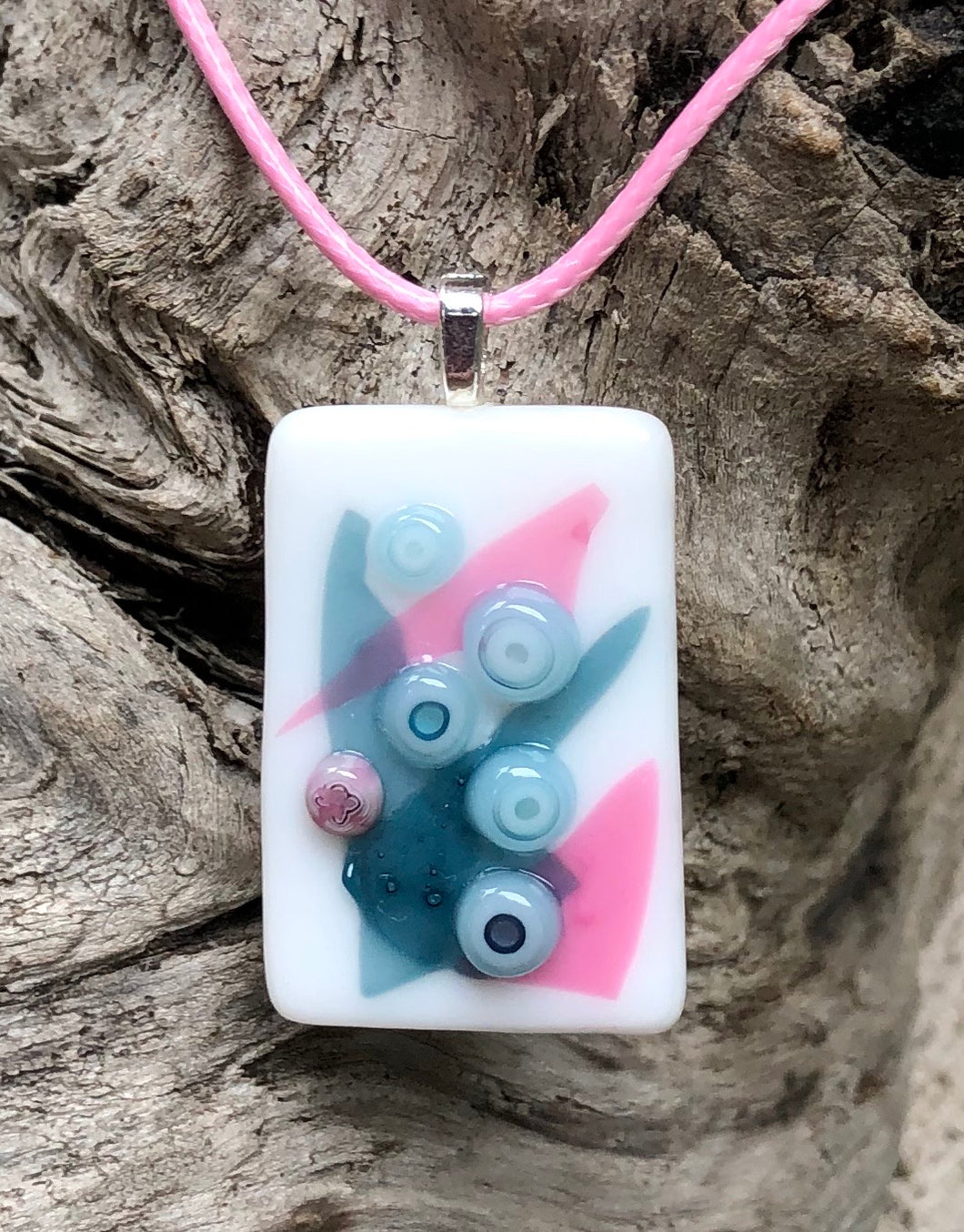 This simple Fused Glass Pendant incorporates Pink and Slate Blue Murrini blooms on Pink and Slate Blue abstract shapes and measures 1 1/4” by 3/4”. The Pale Pink waxed Irish cotton cord is adjustable from 17 1/2” by 18 3/4”.
