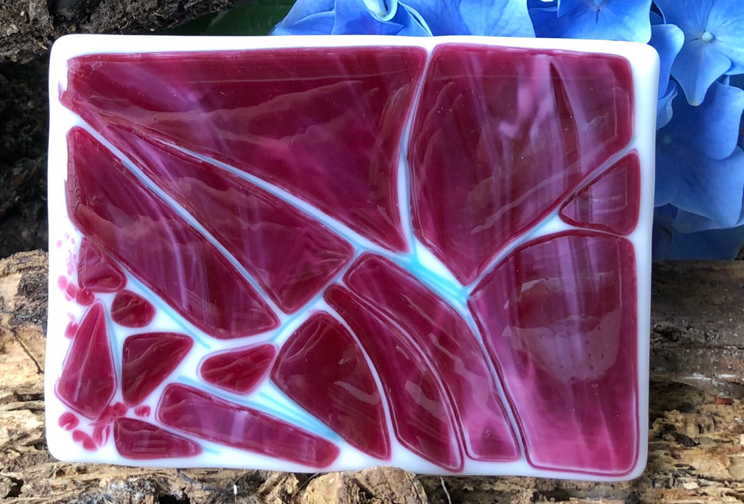 In this Fused Glass dish, geometric slivers of opalescent cranberry glass float on a white background, webbed with light blue slivers.  This dish lays flat with a rippled surface and measures 3 1/2